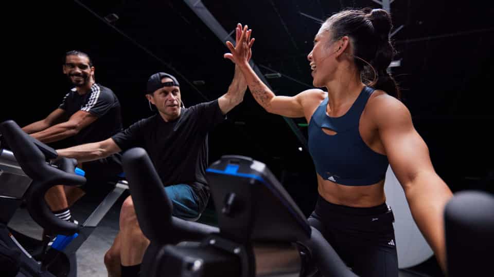Three people on indoor bikes in a dark studio. One in a navy sports bra, others in black tops and shorts, giving a high-five during workout.