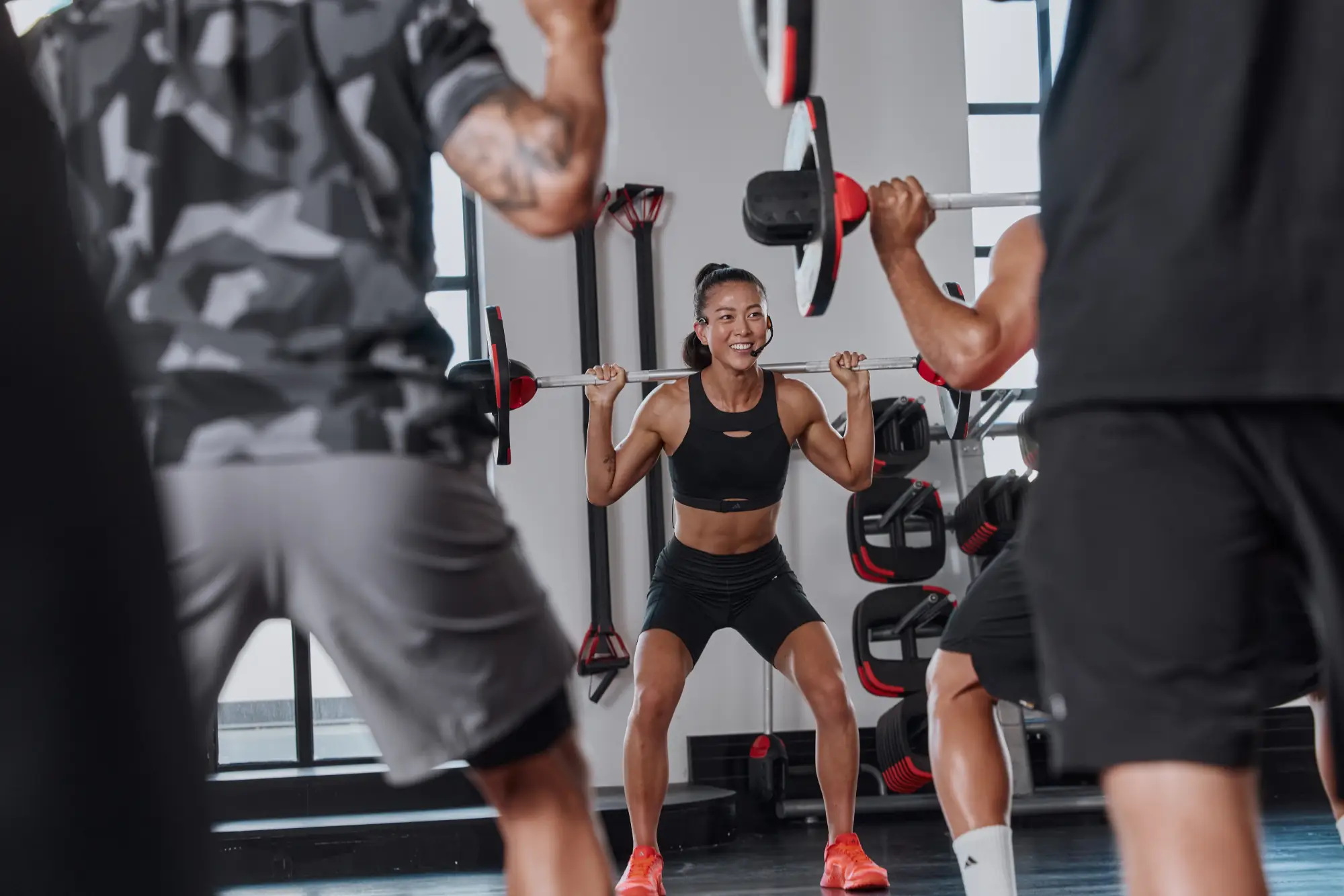 Three people lift barbells with red and black plates in a gym. The central person wears a black sports bra, black shorts, and orange shoes; the others wear dark athletic clothing.