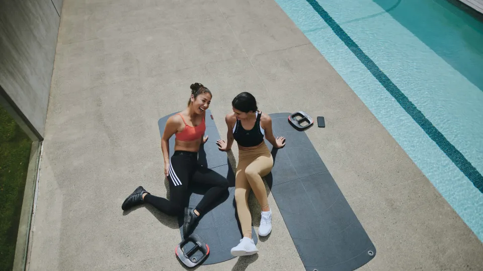 Two Gen Z girls with weight plates and yoga mats discussing workouts poolside