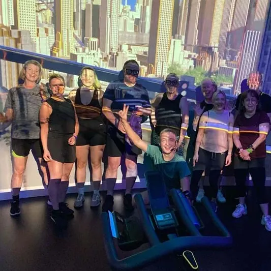 Ten people in athletic wear pose in a dimly lit gym with blue and purple lighting and a cityscape backdrop. One person sits on a machine making a peace sign.