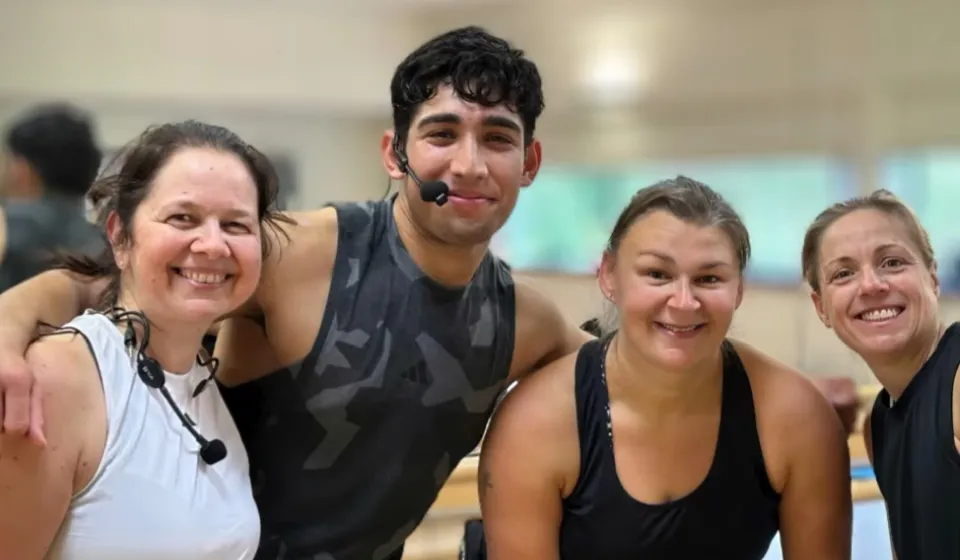 Four people in a gym with light-colored walls and mirrors, wearing workout attire—two in white tops and two in black tops - standing closely together.