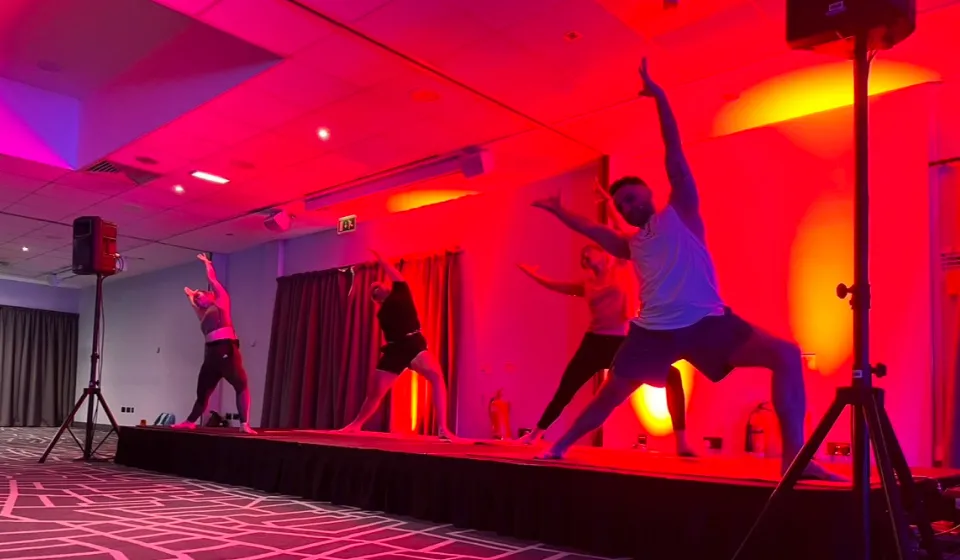 Four people perform yoga poses on a stage with red and orange lighting, patterned carpet, and grey curtains in the background.