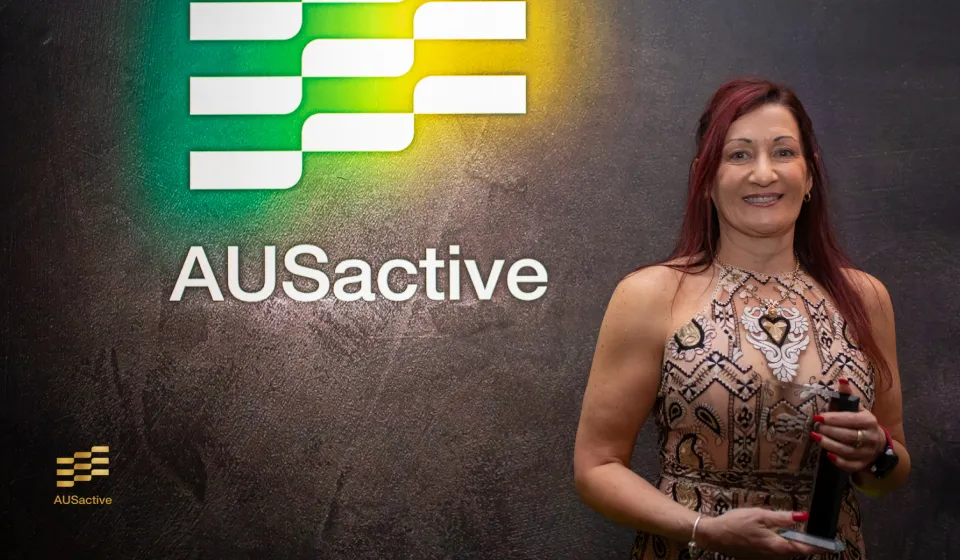 One woman with long red hair in a patterned sleeveless dress holds an award in front of a dark wall with the white and green AUSActive logo and "AUSActive" text in white.