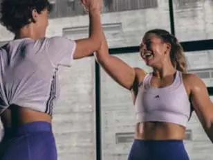 Two people in a bright indoor space high-five each other. One wears a white crop top and blue leggings, the other a white top with blue bottoms. Large windows and gray walls in the background.