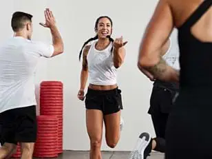  Three people in a gym setting wearing white tops and black shorts. One person runs toward another for a high-five, with red stackable steps visible in the background.
