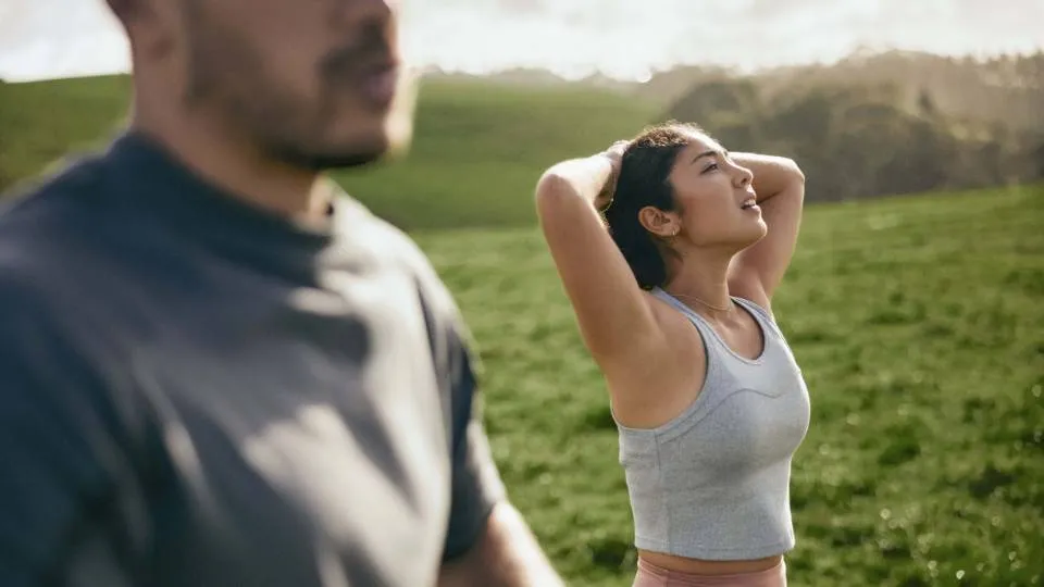 Two people outdoors on green grass with hills in the background. One wears a grey T-shirt, the other a light grey sports top with arms raised behind the head under bright natural light.