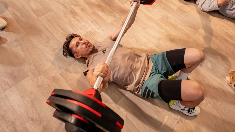Person lying on a wooden floor lifting a barbell with red and black plates, wearing a beige top, green shorts, and black knee sleeves.