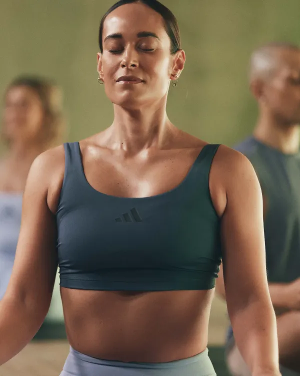 One person in a teal sports bra and light blue leggings standing indoors in a yoga pose. Two others in muted workout gear are visible in the background.