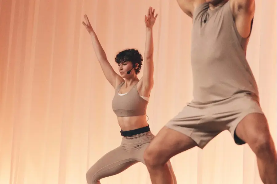 Two people in beige activewear performing a wide-legged stance with arms raised. Soft peach background with vertical drapes creates a warm, minimal studio setting.