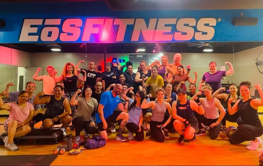 Group of about 24 people posing in a gym with wooden flooring under bright lights. Large blue sign with white and pink text reads “EōS FITNESS” above mirrored wall. Dumbbells visible on floor.